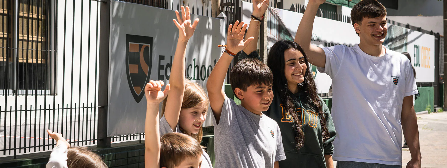 Alumnos de EDU School de distintas edades saludando con la mano frente al cartel del instituto.