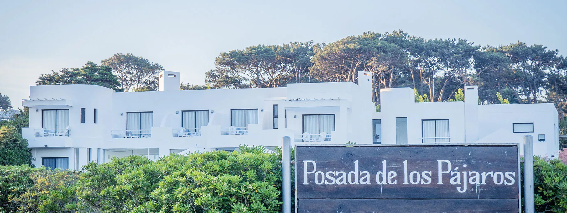 La fachada blanca de la Posada de los Pájaros entre pinos, con su cartel de madera.